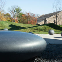 A large ovoid stone sculpture in the foreground with round smaller ball shapes in an outdoor setting.