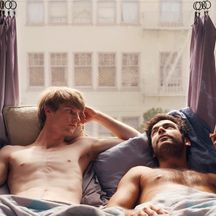 A light-skinned shirtless man reclines and looks at a medium-skinned man, next to him in bed