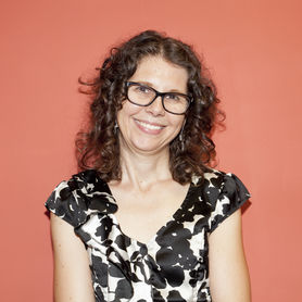 A light skinned woman with glasses and light brown curly hair smiles. Background is bright orange