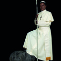A dark skinned man poses dressed like a pope in white robes, holding a staff.