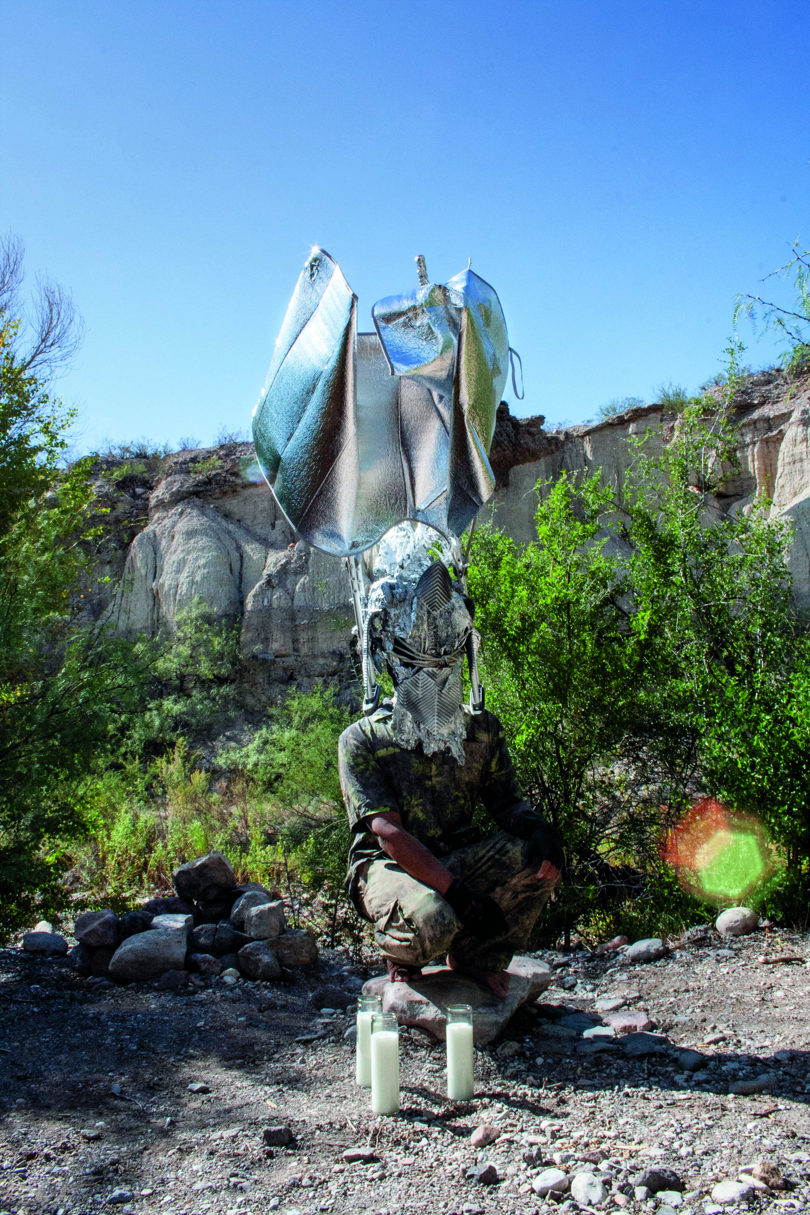 A person in a landscape, crouched down with a reflective silver headdress covering the face and extending high over the head.