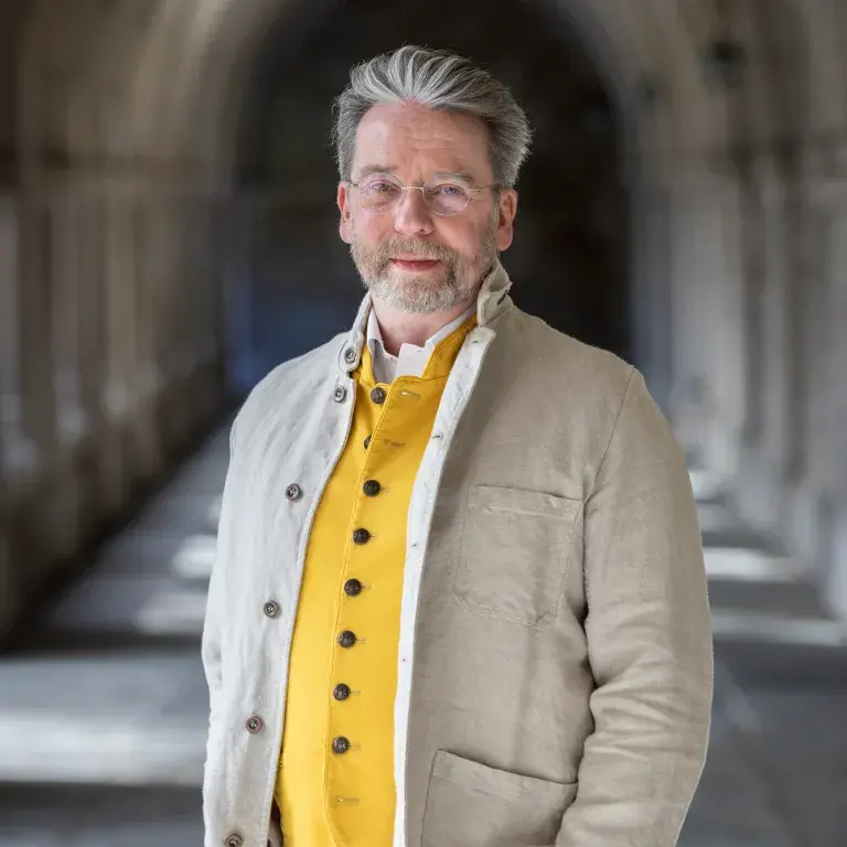 Portrait of a man outside in an arched walkway, wearing a yellow vest and beige jacket.