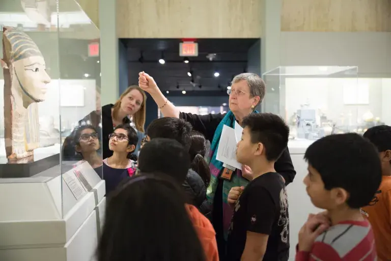A docent points to an Egyptian mask to a group of young students