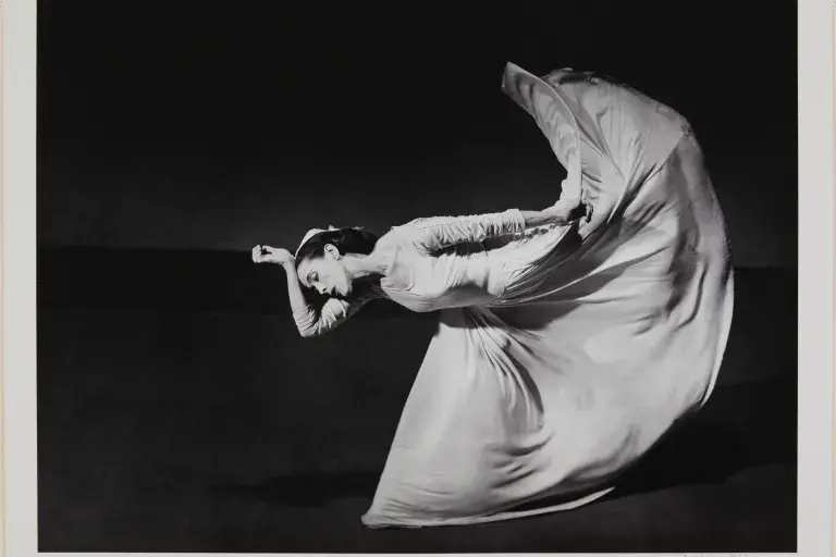 Black and white photograph of a woman in a long white dress dancing.