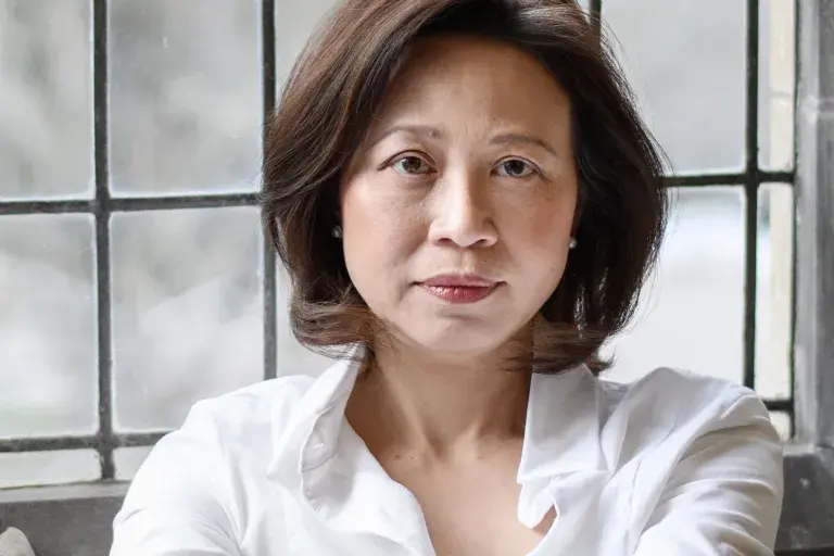 Anne Cheng sitting on a backwards turned chair with her hands crossed in front of a window.