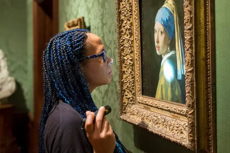 A woman stands close and inspects a painting of a woman with a headscarf and pearl earring