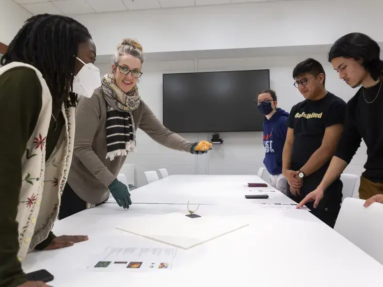 A woman shows a small artifact to a group of students around a table.